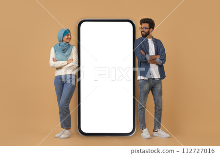 Happy young muslim couple leaning on giant smartphone with blank screen Happy young muslim couple leaning on giant smartphone with blank screen 112727016