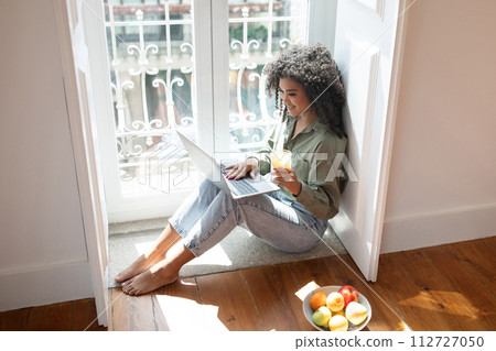 Happy young woman using laptop computer enjoying juice at home 112727050