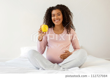Happy young black pregnant woman holding apple while sitting on white bed 112727084