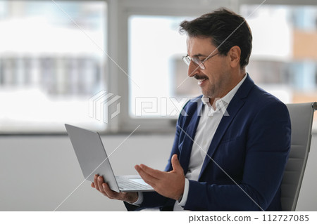 Engaged businessman in a blue suit smiling and gesturing while looking at his laptop Engaged businessman in a blue suit smiling and gesturing while looking at his laptop 112727085