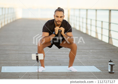 fitness blogger guy squats with dumbbells on a sea pier 112727127