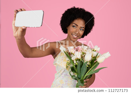 Charming young woman with curly hair taking a selfie, holding a smartphone and a bouquet 112727173