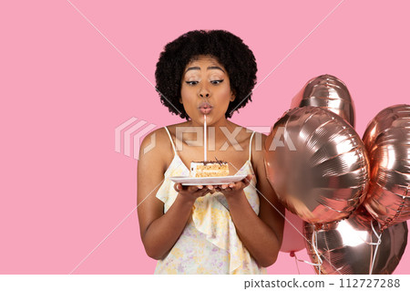 Delighted African American woman with curly hair blowing out a candle on a slice of birthday cake 112727288
