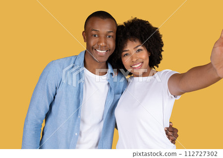 A joyful young couple taking a selfie, with the woman holding the camera and the man embracing her from behind 112727302