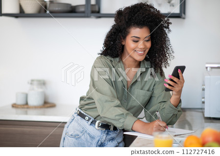 latin young woman stands holding smartphone taking notes in kitchen 112727416