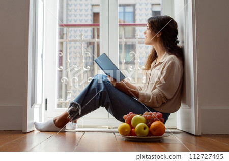 Serene young woman reading book beside bright window at home Serene young woman reading book beside bright window at home 112727495