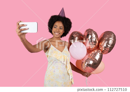 Vibrant African American woman with curly hair and a party hat taking a selfie 112727510