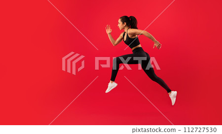 Fit woman in sportswear leaps energetically against vibrant red background Fit woman in sportswear leaps energetically against vibrant red background 112727530