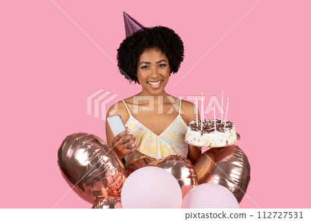 Beaming with happiness, a young woman in a party hat holds a smartphone in one hand and a birthday cake Beaming with happiness, a young woman in a party hat holds a smartphone in one hand and a birthday cake 112727531