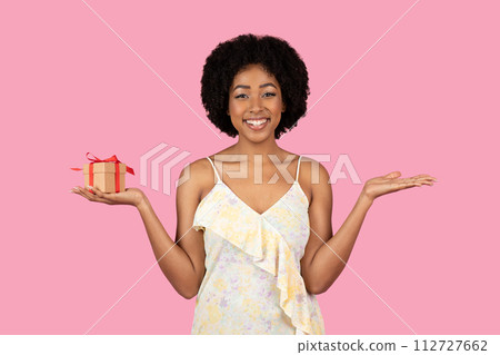 Radiant African American woman with curly hair, smiling and balancing a small gift box with a red ribbon 112727662