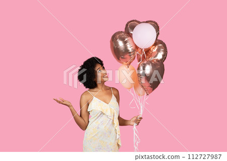 Cheerful African American woman with curly hair, looking up at rose gold and white balloons in her hand 112727987