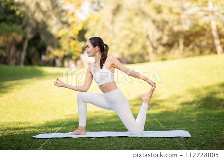 A focused young woman in a white yoga outfit holds an elegant dancer's pose on a mat in a sunny A focused young woman in a white yoga outfit holds an elegant dancer's pose on a mat in a sunny 112728003