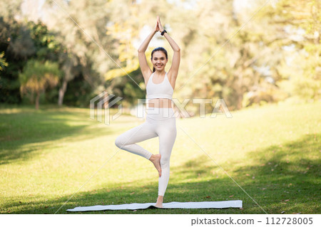 A joyful woman practices the tree pose in a yoga session at a lush park, exemplifying health 112728005