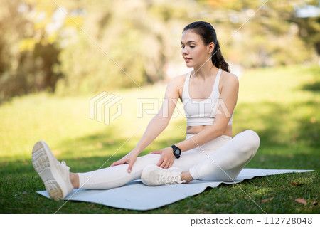 A woman in white athletic wear gently stretches on a yoga mat in the park A woman in white athletic wear gently stretches on a yoga mat in the park 112728048