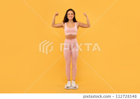 Proud Young Fitness Lady Standing On Scales Showing Biceps, Studio Proud Young Fitness Lady Standing On Scales Showing Biceps, Studio 112728148
