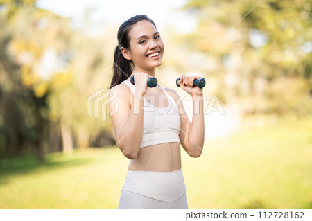 A radiant woman in a white sports bra and leggings holds weights with a confident smile in a sunlit 112728162
