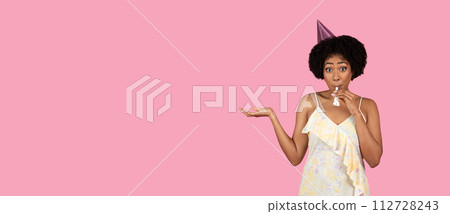 Surprised African American woman with curly hair wearing a party hat and blowing a party horn 112728243