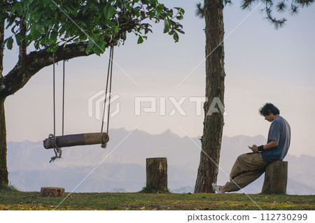 landscape and travel concept with solo freelancer man use tablet work at wooden swing under large tree and layer of mountain landscape and travel concept with solo freelancer man use tablet work at wooden swing under large tree and layer of mountain 112730299