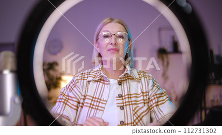 A mature woman blogger is adjusting a ring lamp before streaming online, utilizing modern devices A mature woman blogger is adjusting a ring lamp before streaming online, utilizing modern devices 112731302