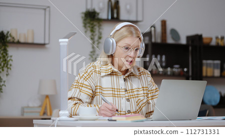 A senior woman is taking a video course, diligently writing down notes while studying online on her laptop 112731331