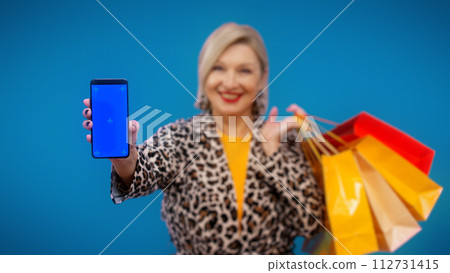 A senior woman is showing her smartphone with a pre-keyed screen and holding shopping bags 112731415