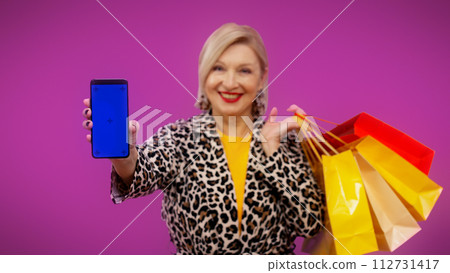 A stylish senior woman is showing a phone with a pre-keyed screen and shopping bags 112731417