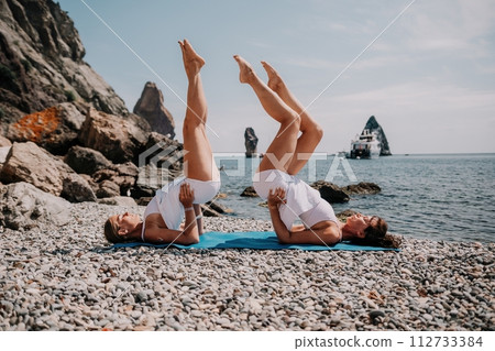 Woman sea yoga. Two Happy women meditating in yoga pose on the beach, ocean and rock mountains. Motivation and inspirational fit and exercising. Healthy lifestyle outdoors in nature, fitness concept. Woman sea yoga. Two Happy women meditating in yoga pose on the beach, ocean and rock mountains. Motivation and inspirational fit and exercising. Healthy lifestyle outdoors in nature, fitness concept. 112733384