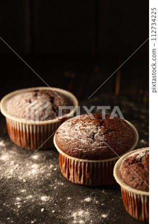 Chocolate muffins with powdered sugar on a black background. Still life close up. Dark moody. Food photo. Chocolate muffins with powdered sugar on a black background. Still life close up. Dark moody. Food photo. 112734225