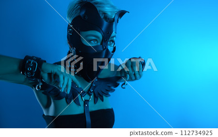 portrait of a hot sexy girl in a leather cat mask and muzzled in bdsm handcuffs on neon blue light 112734925