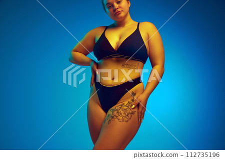 Cropped portrait of female plus-size model posing in dark bikini, underwear in yellow neon light against blue studio background. Cropped portrait of female plus-size model posing in dark bikini, underwear in yellow neon light against blue studio background. 112735196