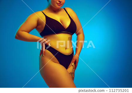 Woman, model plus size in dark lingerie, showcasing natural body curves in yellow neon light against blue studio background. 112735201