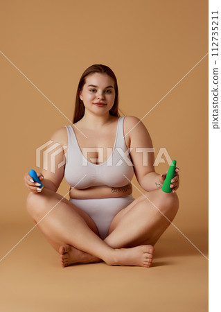 Young overweight woman sitting in lotus pose and holds toys for adults and looking at camera against sandy color studio background. 112735211