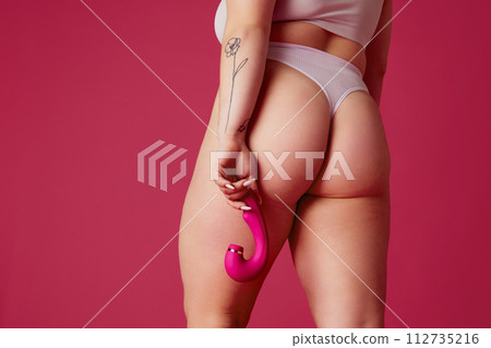 Cropped rear-view portrait of overweight woman in lingerie holds toy for adults near hip against vibrant pink studio background. 112735216