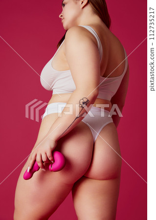 Cropped rear-view portrait of overweight woman in lingerie holds toy for adults behind back against vibrant pink studio background. 112735217