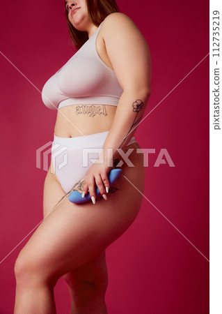 Cropped portrait of young overweight woman in inner wear holds in hands blue toy for adults against vibrant pink studio background. Cropped portrait of young overweight woman in inner wear holds in hands blue toy for adults against vibrant pink studio background. 112735219