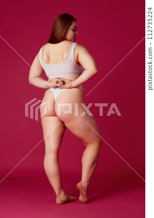 Full length rear-view portrait of young overweight woman posing in lingerie, black bikini showing buttocks, back against pink studio background. Full length rear-view portrait of young overweight woman posing in lingerie, black bikini showing buttocks, back against pink studio background. 112735224