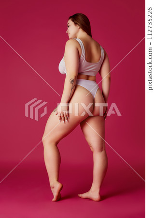 Full length rear-view portrait of young chubby woman posing in lingerie, showing natural curves against grey studio background. Full length rear-view portrait of young chubby woman posing in lingerie, showing natural curves against grey studio background. 112735226