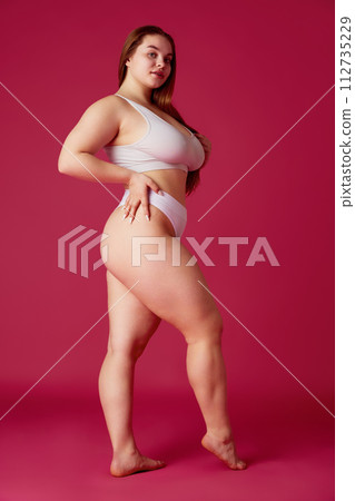 Young, beautiful woman, model plus size posing in lingerie against pink studio background. Overweight girl looks confident. Young, beautiful woman, model plus size posing in lingerie against pink studio background. Overweight girl looks confident. 112735229