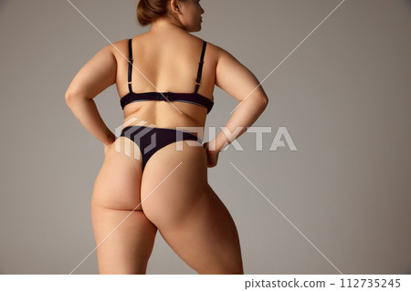 Back view portrait of young attractive woman holds hands on hips and demonstrate her curves against grey studio background. 112735245