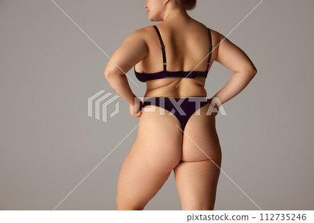 Rear view portrait of model plus size posing holds hands on hips showing her natural body curves against grey studio background. Rear view portrait of model plus size posing holds hands on hips showing her natural body curves against grey studio background. 112735246