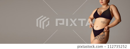 Banner. Cropped portrait of female plus-size model posing in dark bikini against grey studio background with negative space. Banner. Cropped portrait of female plus-size model posing in dark bikini against grey studio background with negative space. 112735252