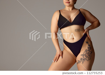 Cropped portrait of female plus-size model posing in dark bikini, underwear against grey studio background. Copy space. Cropped portrait of female plus-size model posing in dark bikini, underwear against grey studio background. Copy space. 112735253