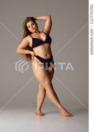 Full-length portrait of woman with tattoo on thigh in dark lingerie posing confidently holds hand on hip against grey studio background. Full-length portrait of woman with tattoo on thigh in dark lingerie posing confidently holds hand on hip against grey studio background. 112735262