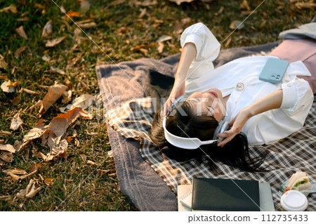 Pleased young woman listening to music with headphones on picnic blanket with closed eyes 112735433