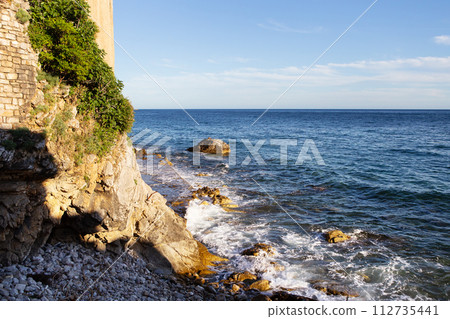 A tranquil seascape with rocky shores, lush greenery, and a clear blue sky. 112735441