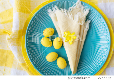 This vibrant Easter table setting features a blue plate adorned with yellow eggs and a delicate flower. The checkered tablecloth adds a festive touch. 112735486