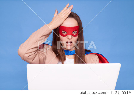 Upset sad woman wearing superhero costume standing isolated over blue background working on laptop having problems with work showing facepalm gesture Upset sad woman wearing superhero costume standing isolated over blue background working on laptop having problems with work showing facepalm gesture 112735519