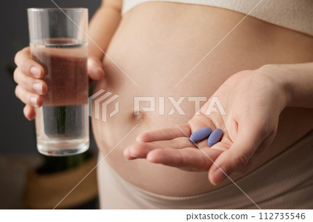 Unrecognizable pregnant woman with bare belly expectant mother touching her tummy holding glass of fresh water and medicine for pregnancy health standing in home interior Unrecognizable pregnant woman with bare belly expectant mother touching her tummy holding glass of fresh water and medicine for pregnancy health standing in home interior 112735546