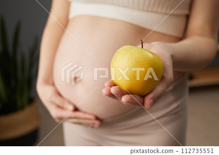 Unknown beautiful pregnant woman with bare belly standing in home interior holding fresh organic yellow apple prefers healthy eating while expecting baby 112735551