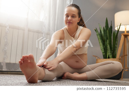 Caucasian pregnant woman wearing beige top and leggings exercising at home stretching her legs enjoying gymnastic exercising in light living room interior 112735595